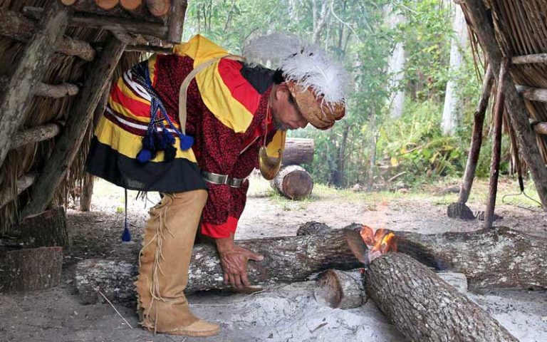 seminole man dressed in ceremonial garments lighting fire in lean-to enclosure with trees at ah-tah-thi-ki museum