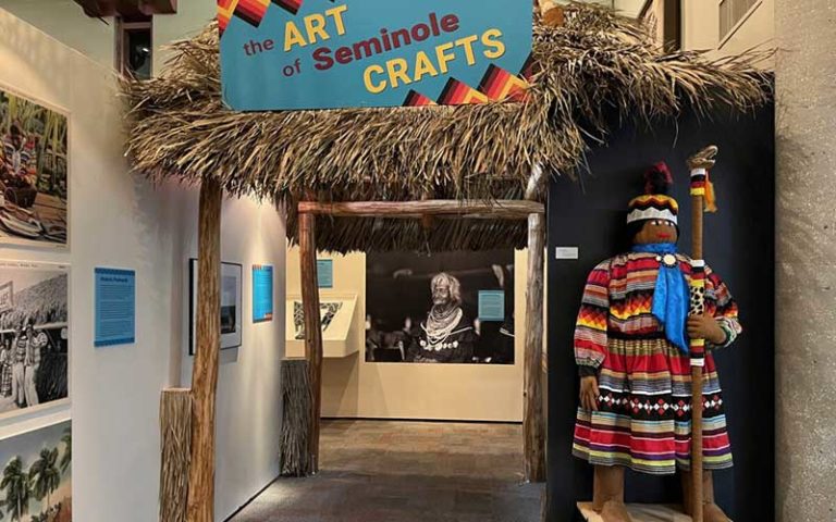 The Art of Seminole Crafts sign with thatched roof structure and images hanging on wall and manekin with garments exhibit at ah-tah-thi-ki museum