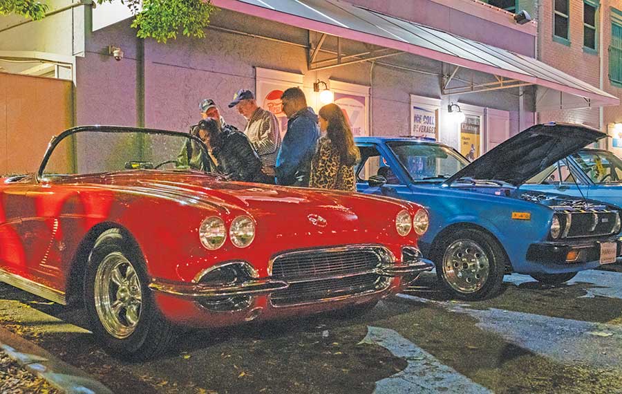 group looks at red classic convertible next to blue car with hood up on street at night at old town car show