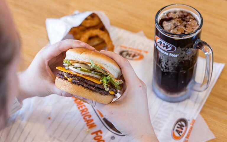 hands holding deluxe burger about to bite with onion rings and mug drink on tabletop at a&w all american food old town