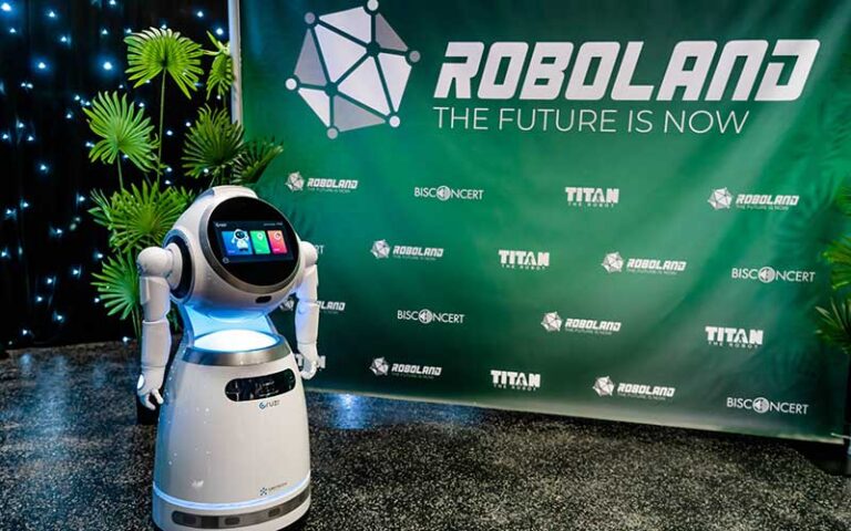 small domestic robot with round touch screen arms and mobility with green roboland banner and plant at roboland orlando