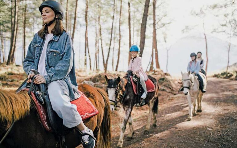 woman horseback rider with helmet followed by kids with helmets riding horses on wooded trail at green lakes farm kissimmee