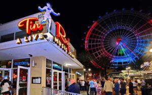 angled front exterior at night with sign crowds and lit up wheel at tin roof orlando icon park