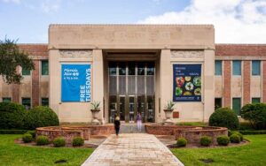 front exterior daytime stone facade with people walking path to door between round walls and shrubs at cummer museum of art and gardens jacksonville