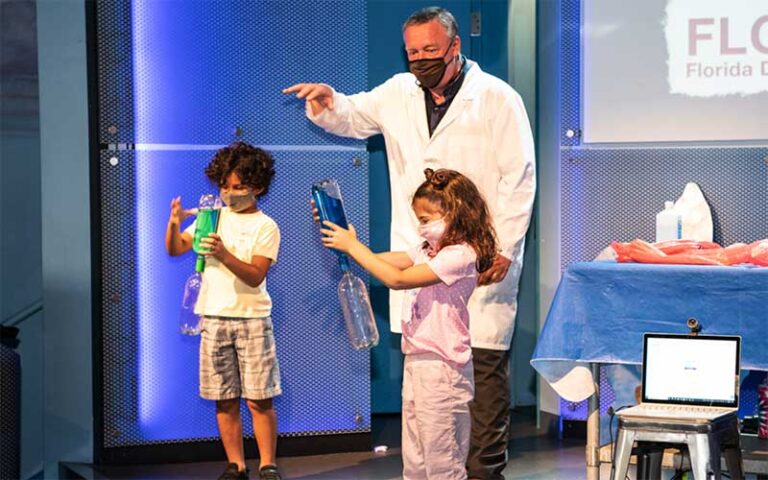 man in lab coat on stage with two kids holding bottles for experiment show at museum of discovery and science ft lauderdale