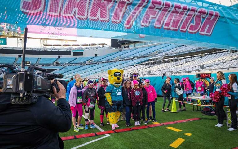 running event with cameras and sports mascot posing with group on finish line in arena at tiaa bank field jacksonville