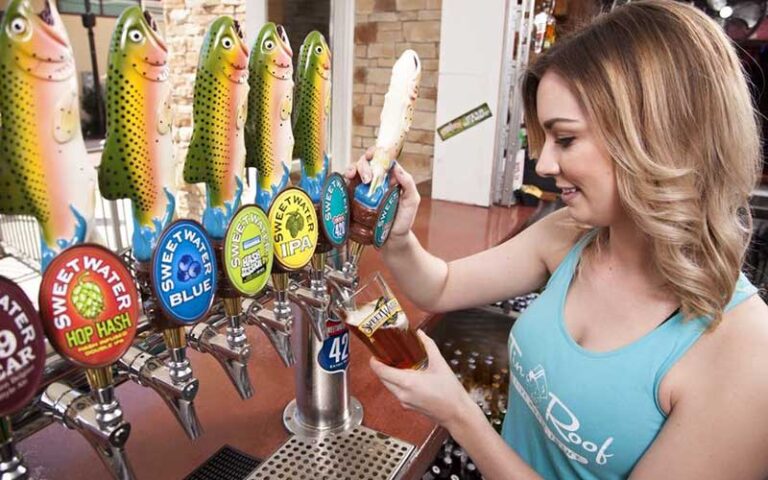 woman server in blue top pouring glass of beer from row of taps with trout handles at tin roof orlando icon park