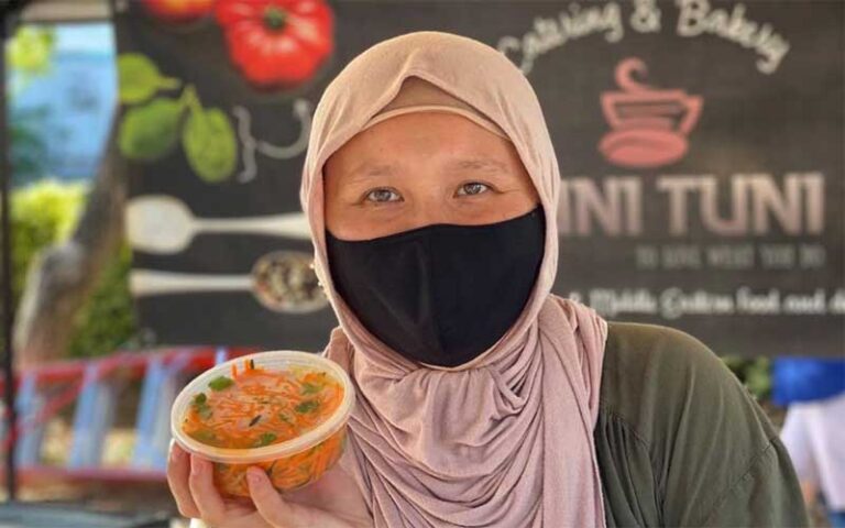 woman in mask holding food container with sign in booth at sarasota farmers market