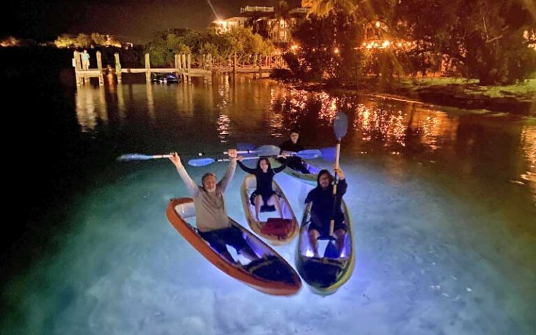 family in illuminated kayaks holding up oars with dock and houses at night kayak key west