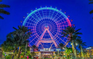 night view of the wheel with blue and red lighting and building with palms at icon park orlando