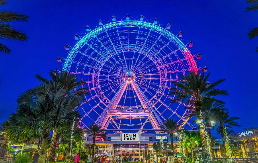 night view of the wheel with blue and red lighting and building with palms at icon park orlando