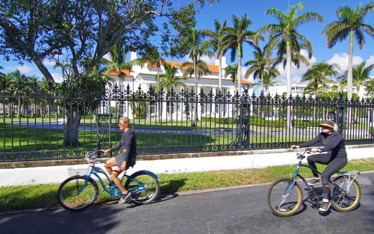 bicycle riders on path passing iron fence with white stately home at palm beach lake trail west palm beach