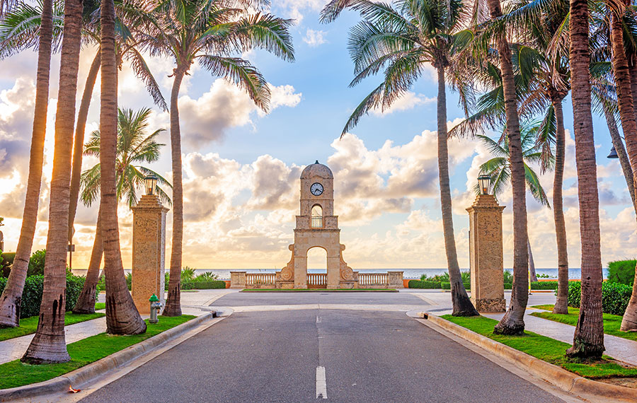 view of end of worth avenue with centered clock tower blue sky sunrise and palm trees
