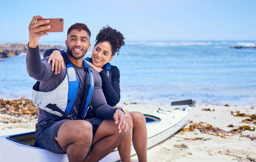 couple smiling for mobile phone selfie seated on kayak on inlet beach