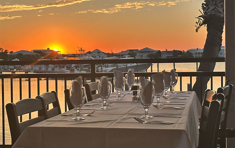 table on patio with dining setting overlooking marina with orange sunset louisiana lagniappe destin