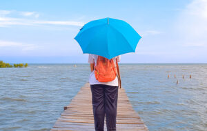 woman with orange backpack holding umbrella on pier overlooking coastal waterway with gray sky