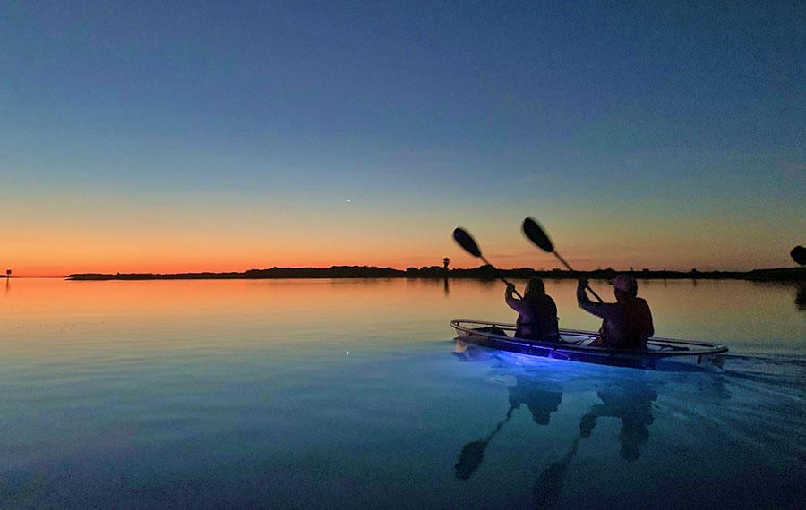 couple in illuminated kayak paddling toward setting sun over dark water at get up and go kayaking ft pierce
