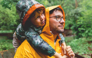father piggybacking young son both wearing wet raincoats in woods