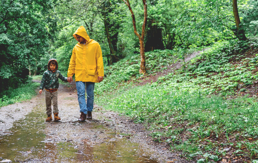 dad and son in raincoats walking along wooded trail with puddles