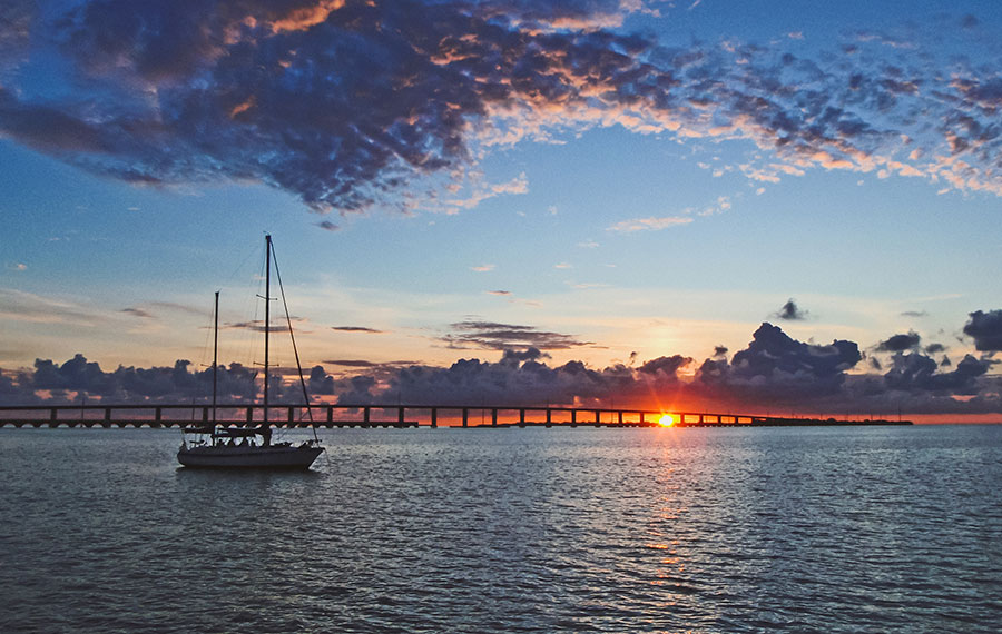 sailboat silhouetted against sunset over bay with bridge and clouds