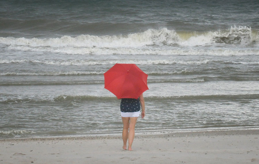 woman carrying red umbrella walking towards storm tossed ocean along beach