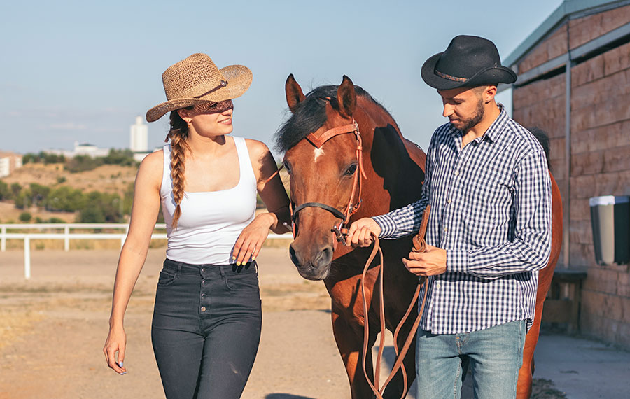 couple wearing western clothes walking with horse at corral