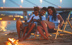 family of three sitting in lounge chairs at dusk with beach fire and lighted bay bridge