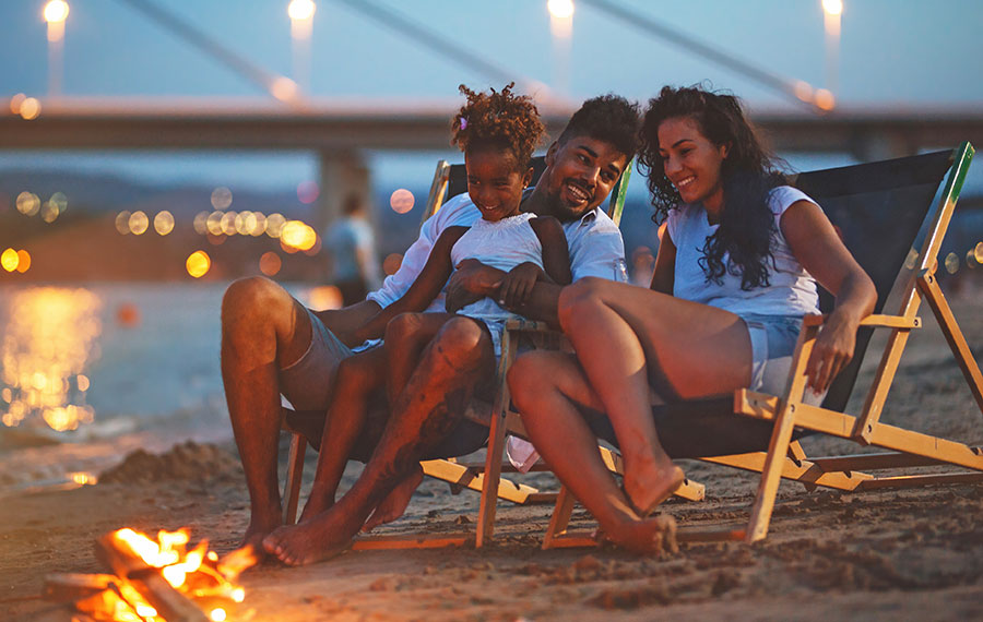 family of three sitting in lounge chairs at dusk with beach fire and lighted bay bridge